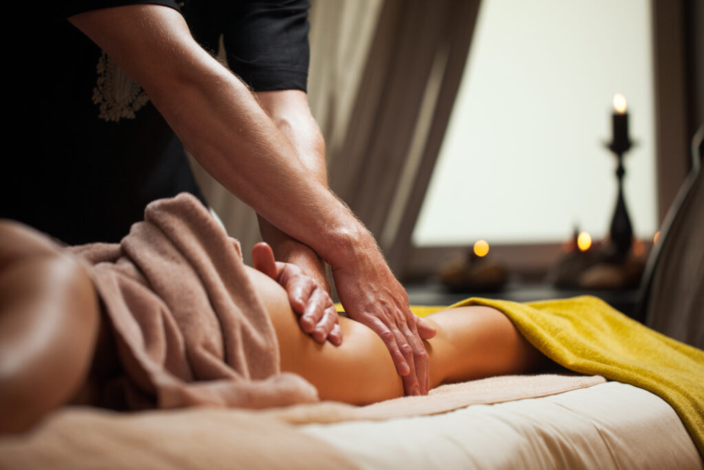 Person erhält eine Beinmassage auf einem Massagetisch, der teilweise mit Handtüchern bedeckt ist. Kerzenlicht im Hintergrund sorgt für eine entspannende Atmosphäre.