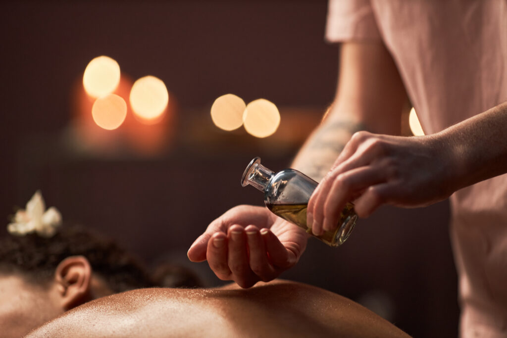 Person gießt Massageöl aus einer kleinen Flasche auf ihre Hand und bereitet sich darauf vor, den Rücken einer anderen liegenden Person zu massieren. Brennende Kerzen sind im Hintergrund verschwommen.